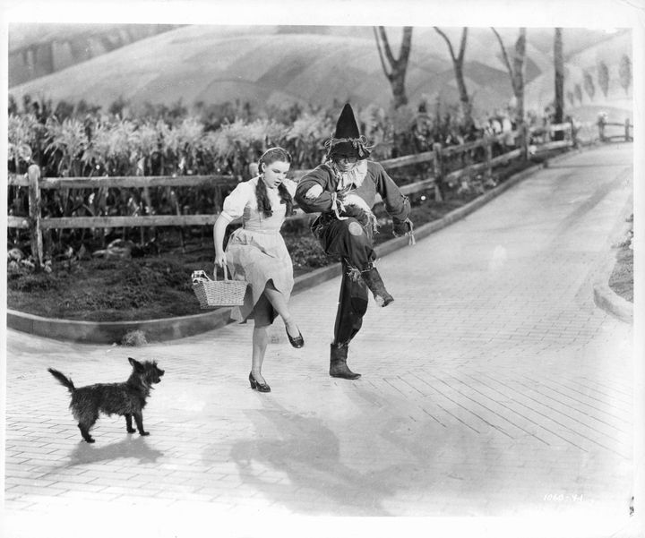 Actors Judy Garland and Ray Bolger dance in character on the 'Yellow Brick Road' as dog dog 'Toto' watches, in a scene from the film 'The Wizard Of Oz', 1939. (Photo by Metro-Goldwyn-Mayer/Getty Images)